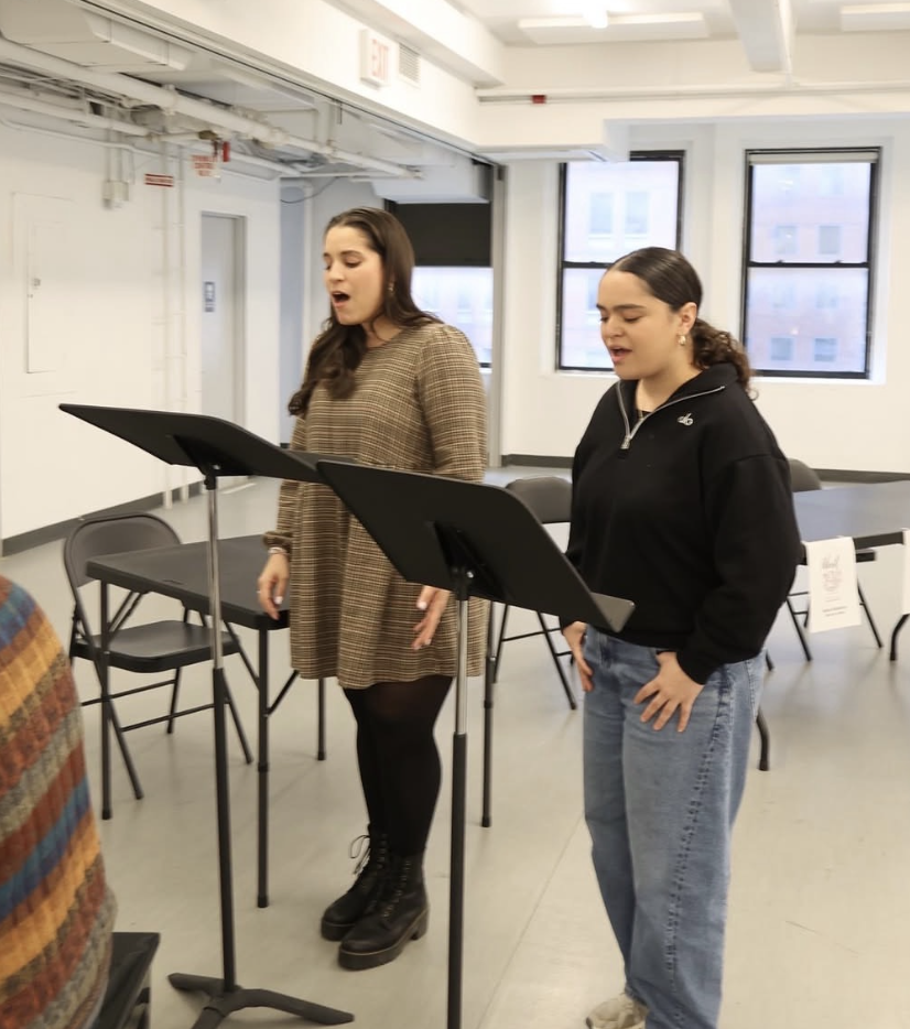 Two young women are singing or speaking while standing at music stands in a bright, modern room with large windows and empty chairs around them.