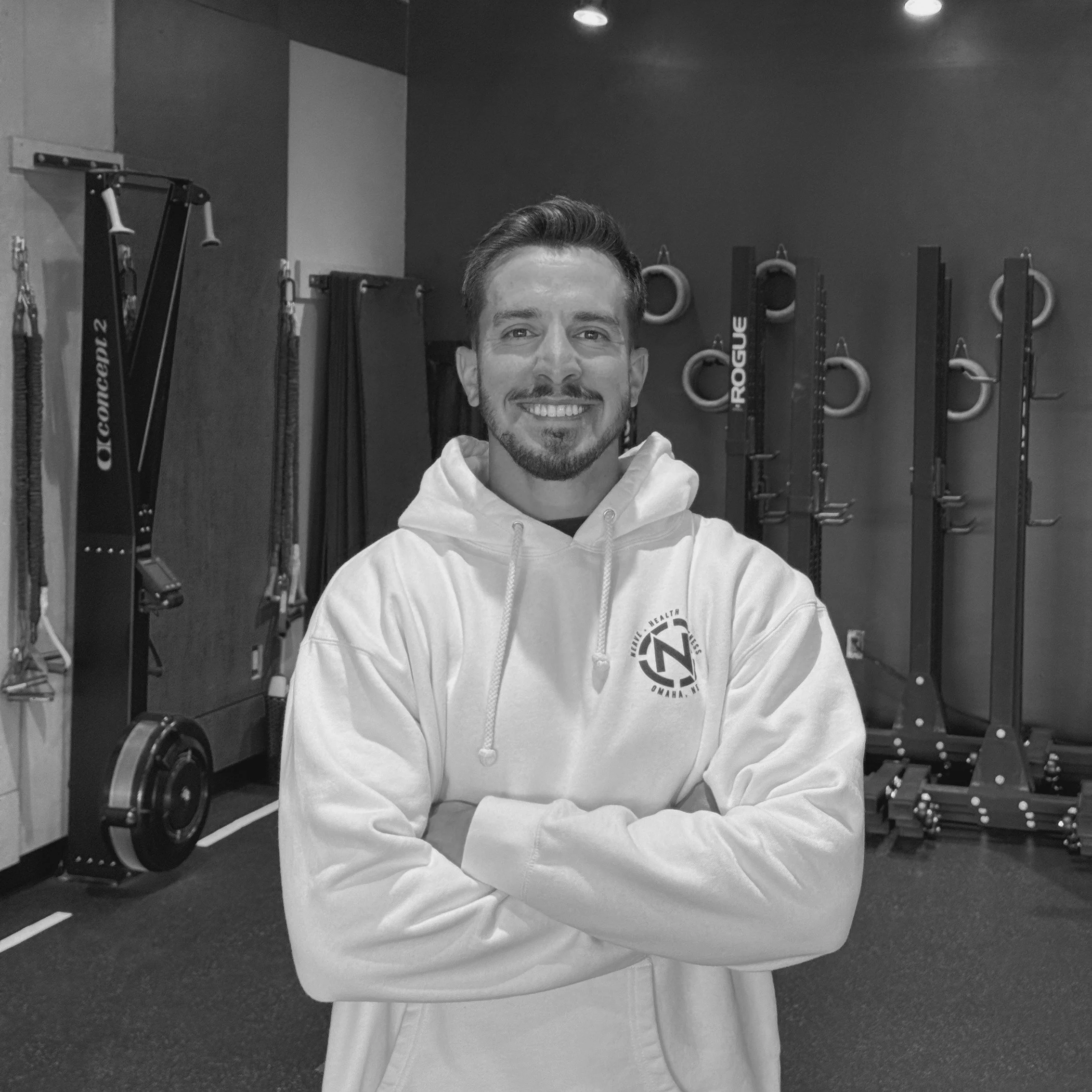 A young man with a beard and short hair smiling at the camera, wearing a hoodie, inside a gym. Behind him, gym equipment like resistance bands, gymnastic rings, and weight plates are visible.