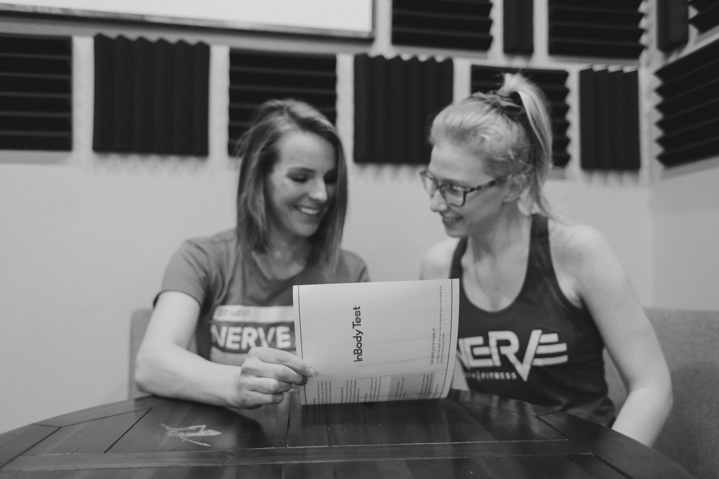 Two women sitting at a table in a recording studio, looking at a piece of paper and smiling.