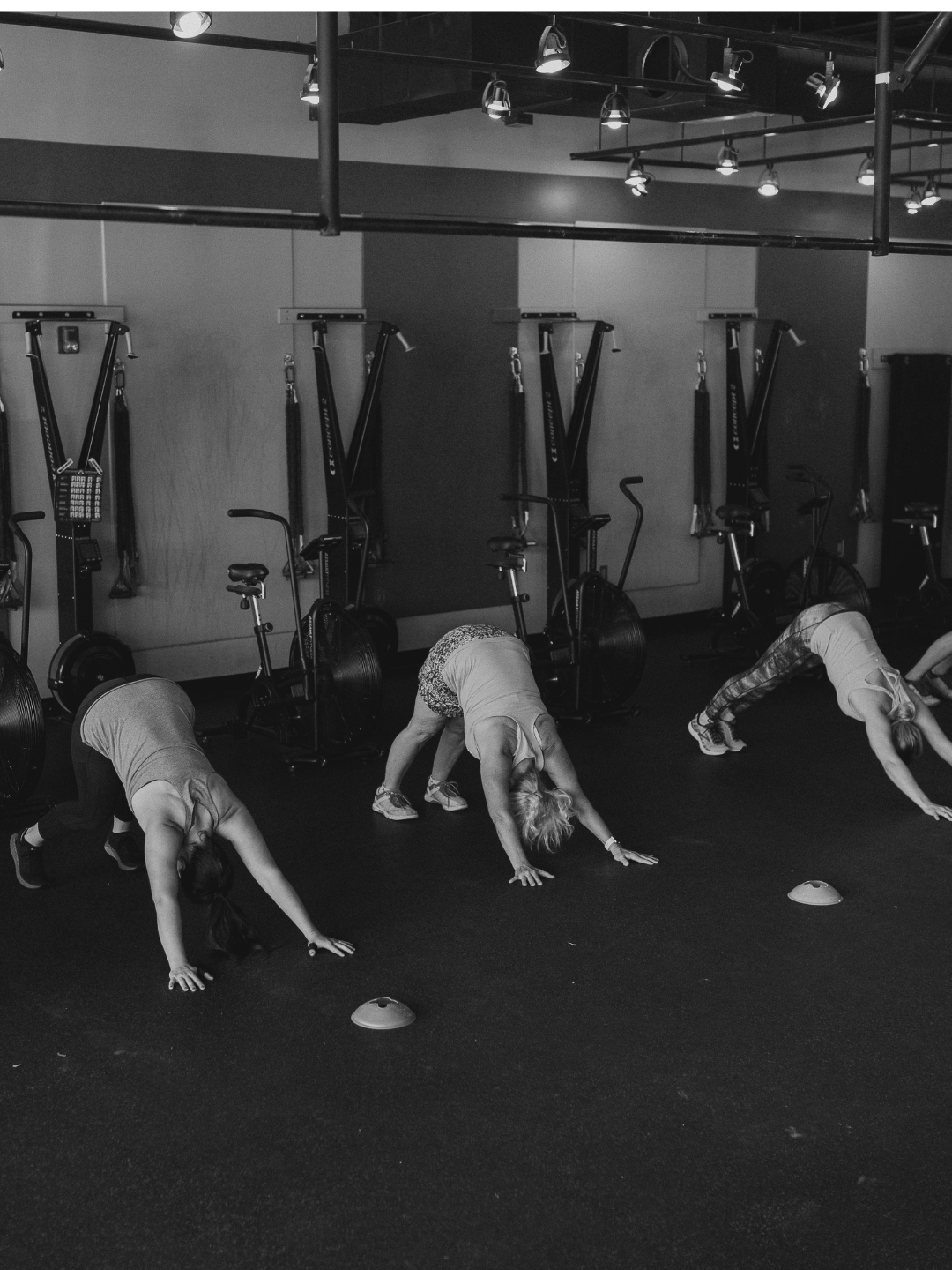 Three women in a gym are doing downward dog yoga poses on a black mat floor. There is gym equipment mounted on the wall in the background, including stationary bikes and rowing machines, with overhead lights above.