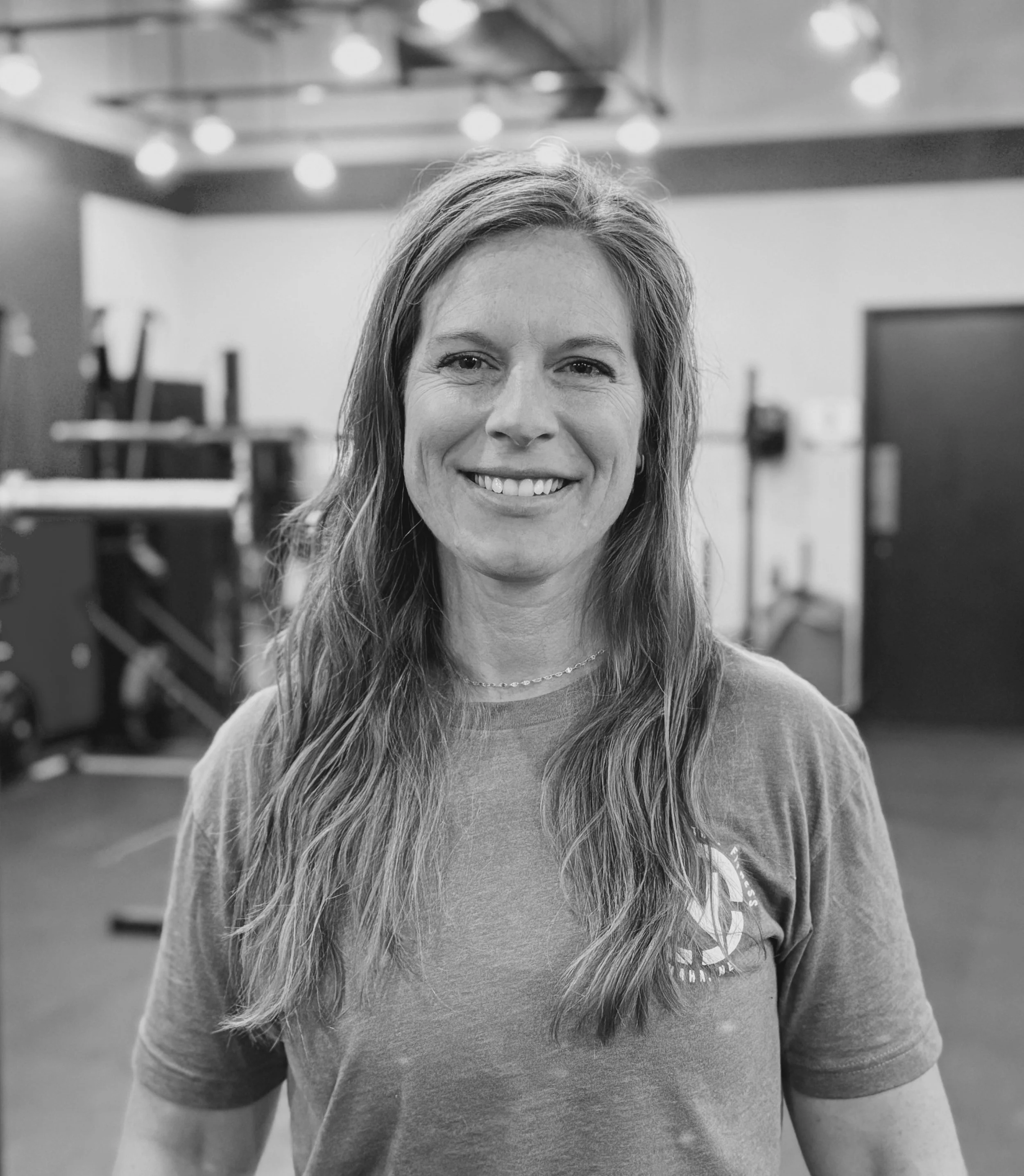 Black and white photo of a woman with long hair smiling in an indoor setting, possibly a gym or studio, with blurred background equipment and lights.