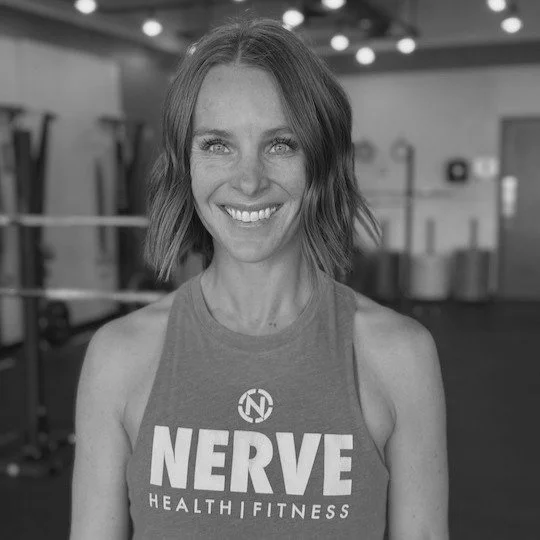 A woman smiling in a gym, wearing a sleeveless top with 'NERVE HEALTH & FITNESS' printed on it.