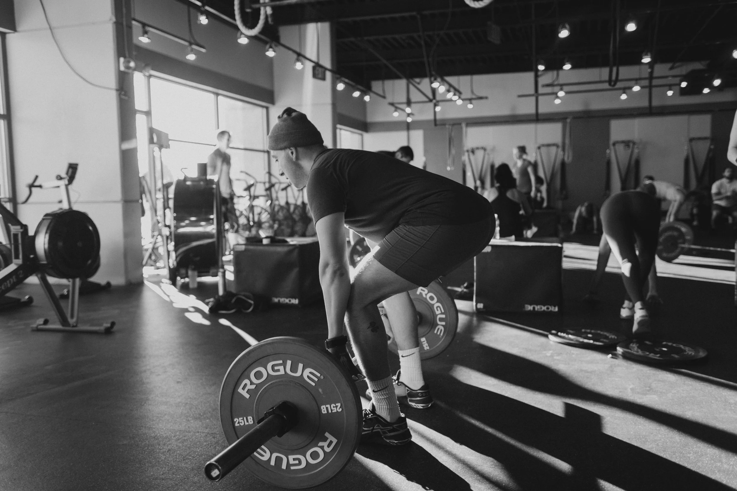 A person wearing a beanie and athletic clothing lifting a barbell with weights in a gym, with several other people working out in the background.