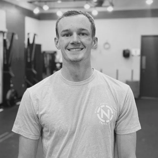 A young man smiling in a gym, wearing a light-colored T-shirt with a logo on the chest.