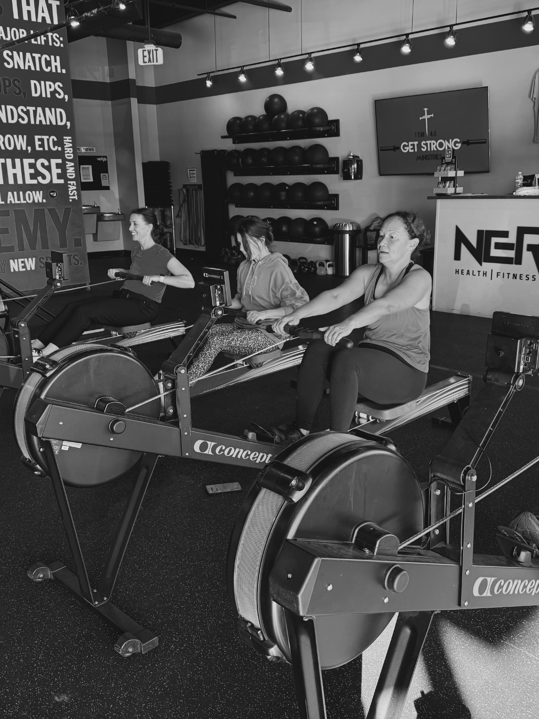 Three women working out on rowing machines in a gym. Gym equipment, kettlebells, and a TV with motivational text are visible.