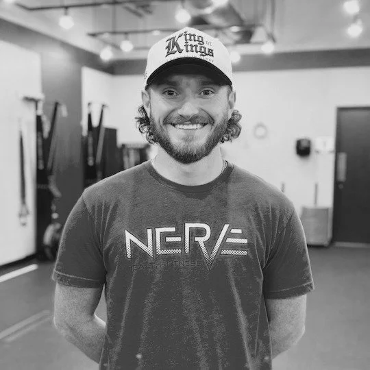A smiling man with curly hair and a beard wearing a cap that says 'King of the Kings' and a t-shirt that says 'NERV' standing in a gym.
