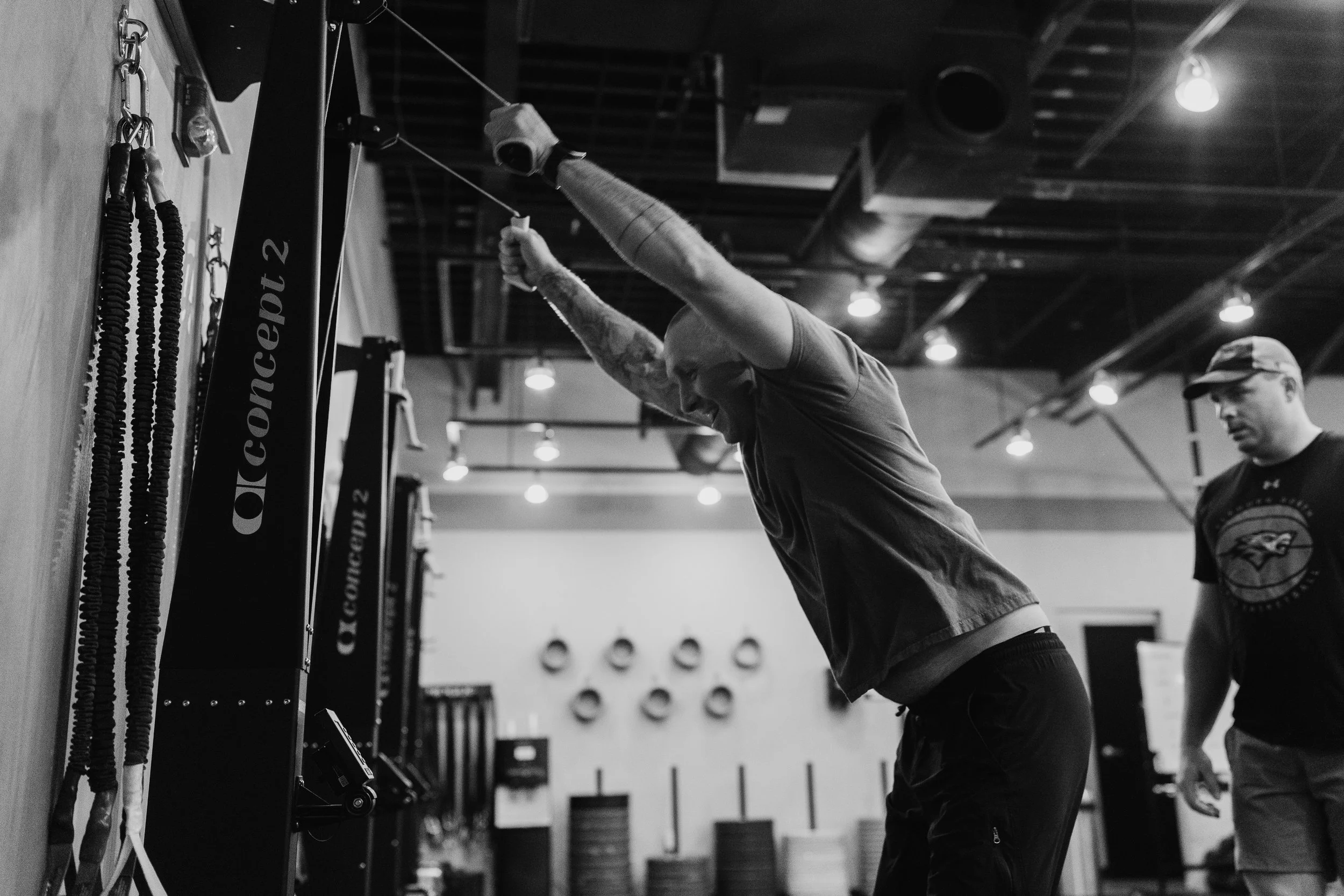 A man in a gym stretching with resistance bands attached to a wall, with another person observing in the background.