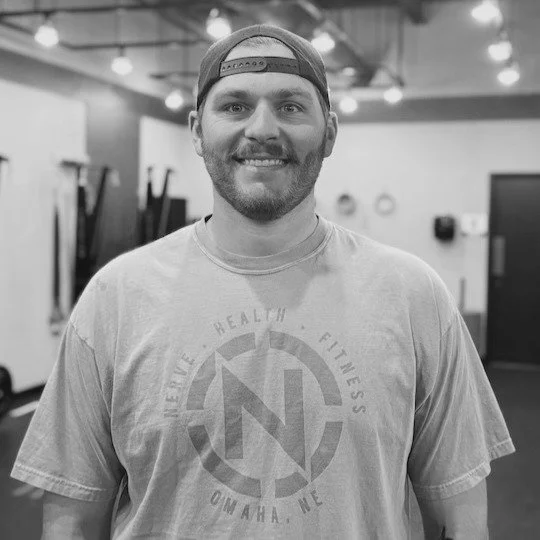 A smiling man with a beard wearing a backwards baseball cap and t-shirt, standing in a gym or fitness center.