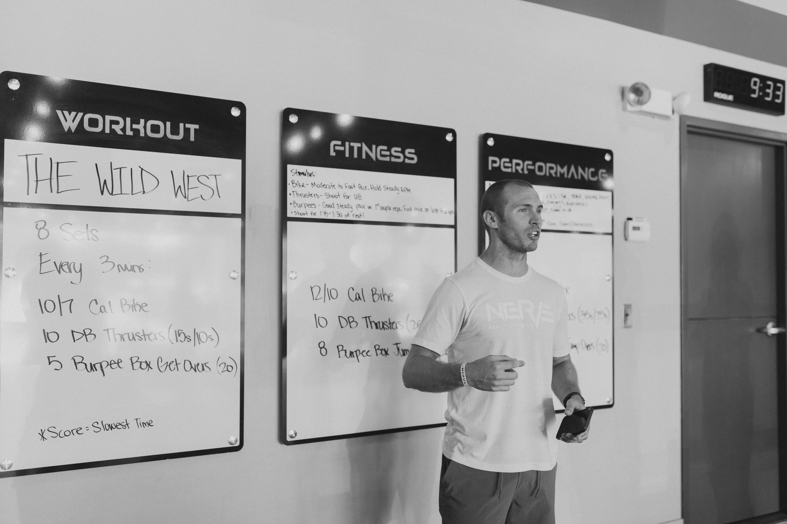 A man in a workout t-shirt holding a phone and standing in front of whiteboards with workout instructions. The signs read "Workout," "Fitness," and "Performance," with handwritten exercise details. An electronic clock displays 9:33.