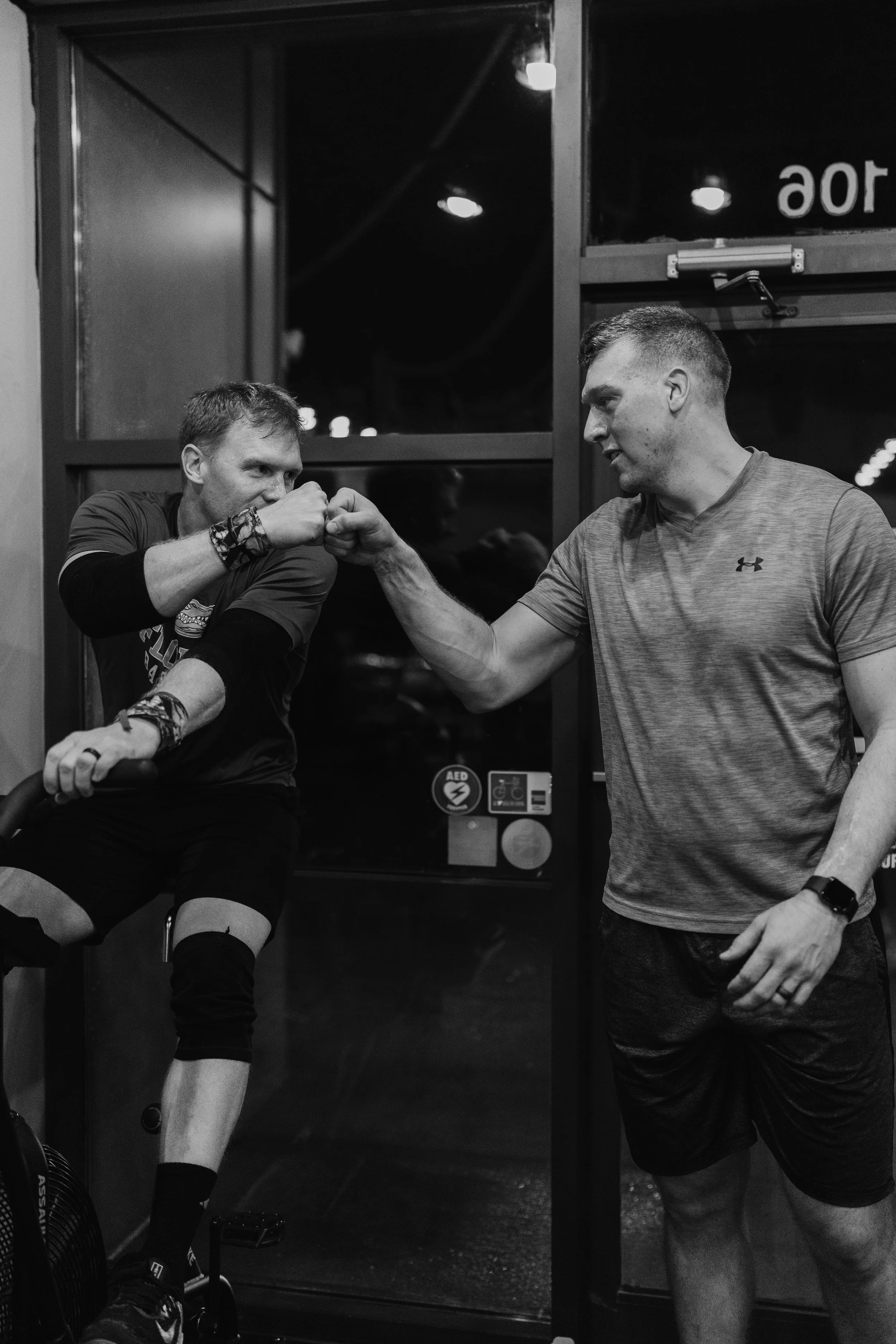 Two men fist bumping in a gym, one of whom is seated on a stationary bike while the other stands next to him. Both are wearing athletic clothing.