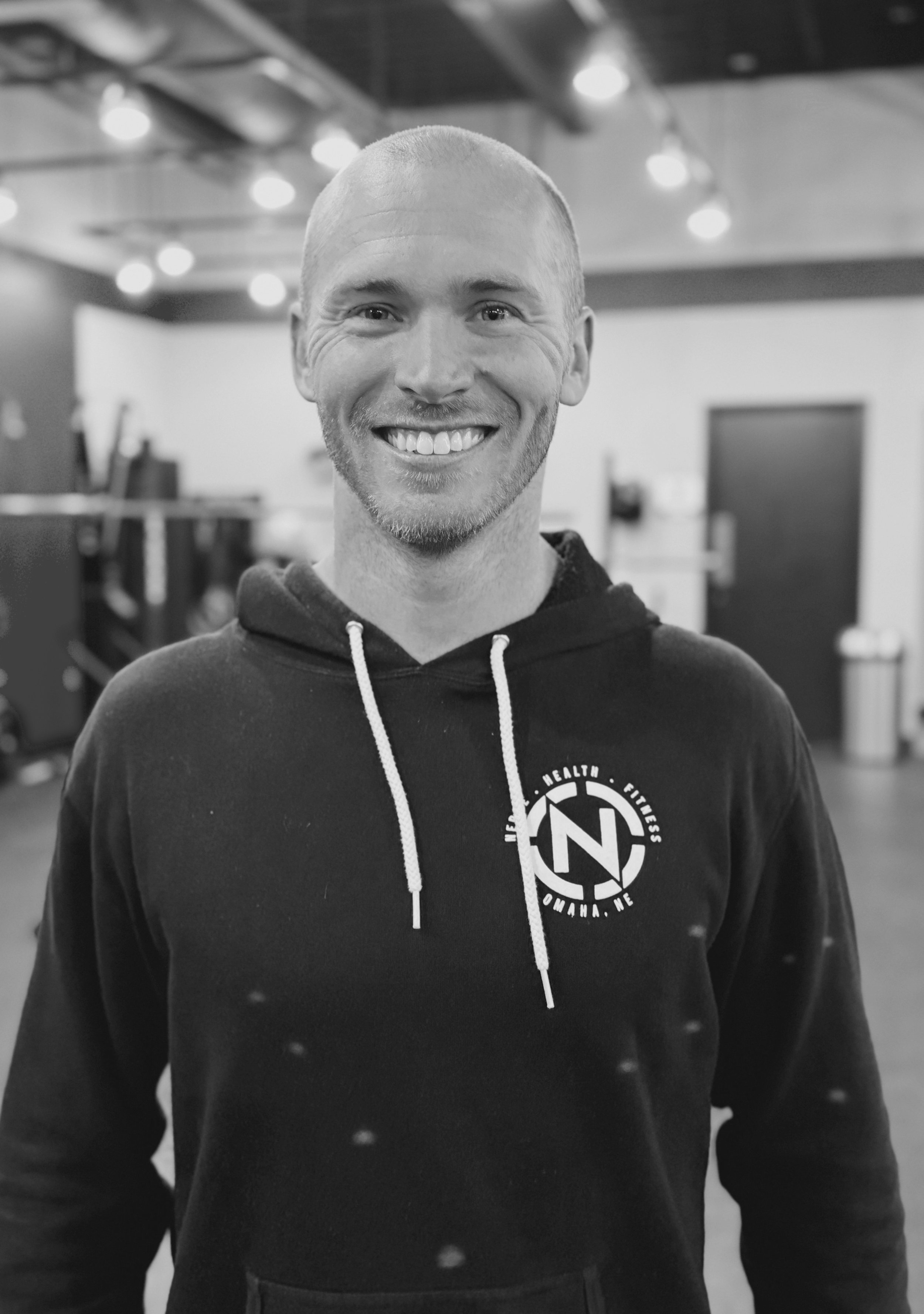 A man with a shaved head and a beard smiling, wearing a black hoodie with a logo on the chest, in a gym or fitness center with gym equipment in the background.