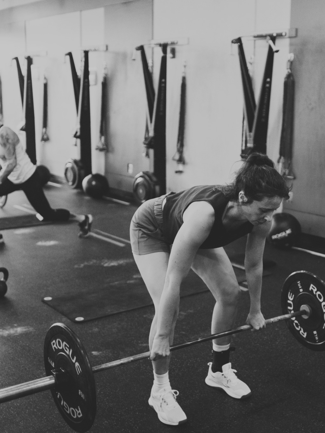 Women lifting a barbell in a gym, with workout equipment and other people exercising in the background.