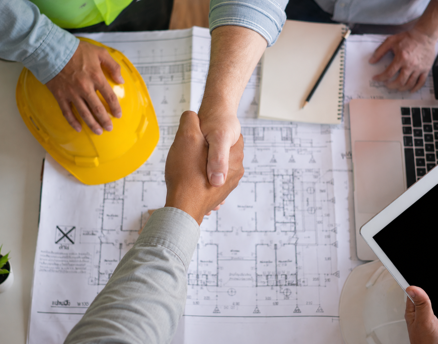 Two people shake hands over an architectural blueprint on a desk. One person is holding a yellow safety helmet, and various office supplies and electronic devices are also on the table.