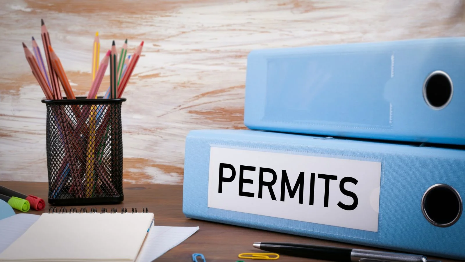 Office desk with a black pen holder filled with colored pencils, a notebook, highlighters, and paper clips, next to two stacked blue folders labeled 'Permits' on a wooden surface with a white and brown wood textured background.