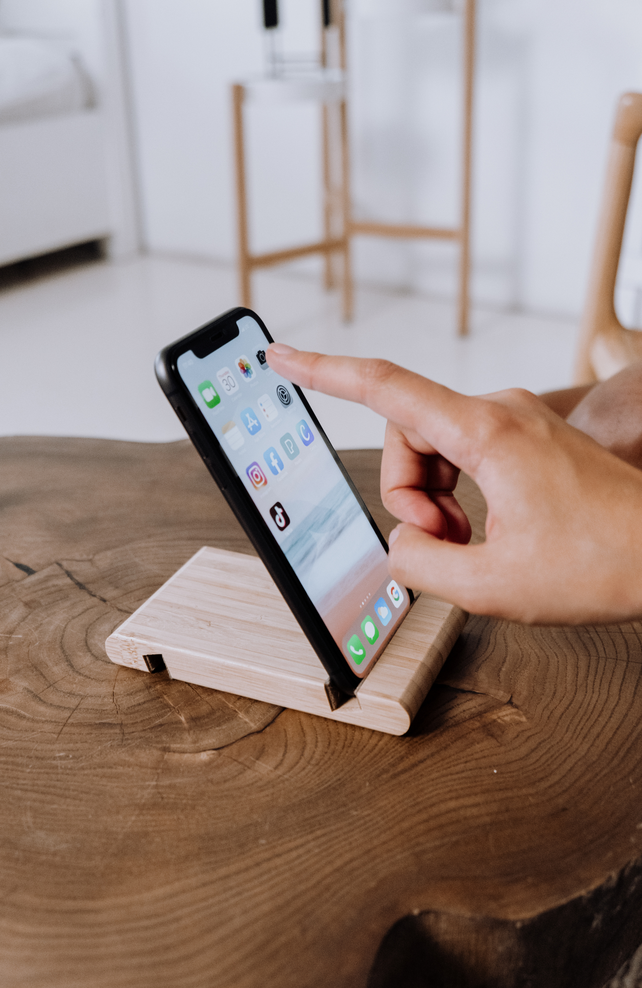 A person holding a smartphone with various app icons on the screen, positioned on a wooden stand on a wooden table.