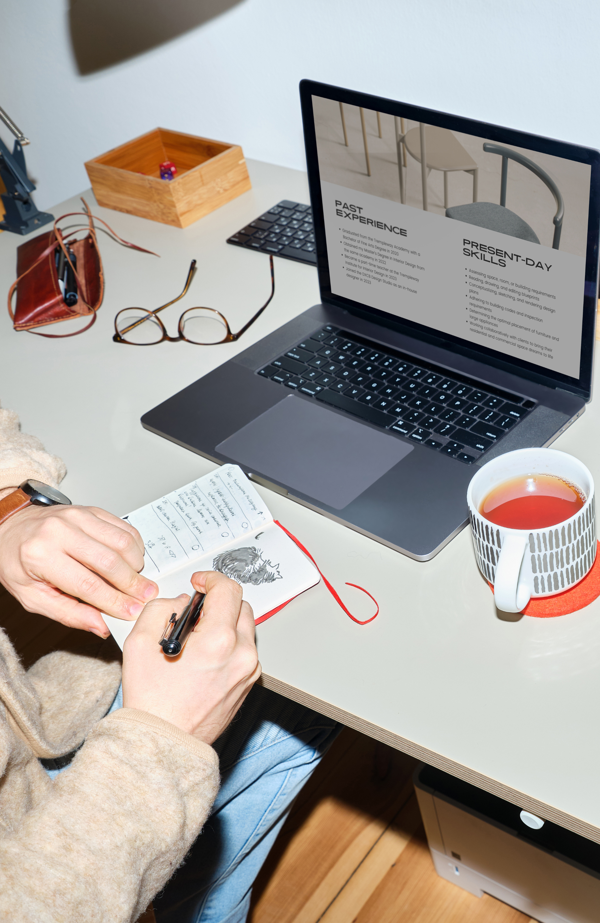 A person sketching a dog in a notebook at a desk with a laptop displaying past and present-day skills, a cup of tea, glasses, and various desk accessories.