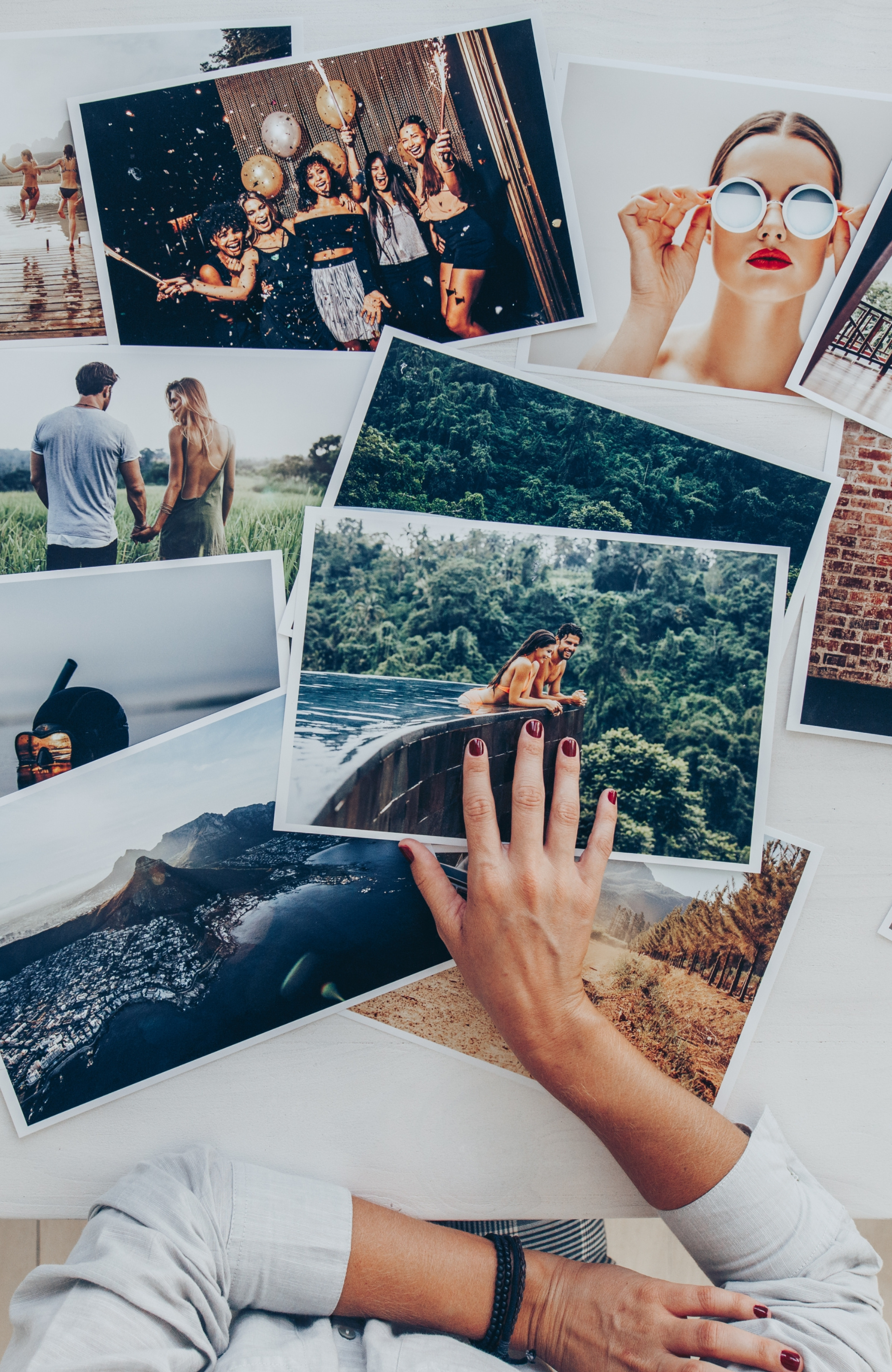 An arrangement of printed photographs on a white surface, depicting various scenes such as people celebrating, a woman wearing sunglasses, couples holding hands, and scenic landscapes like forests, waterfalls, and rocky coastlines.