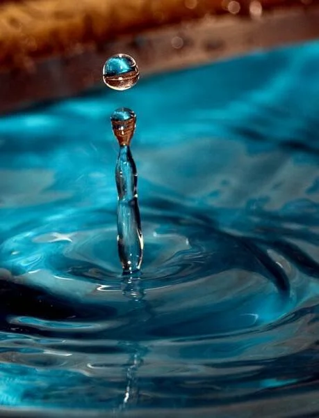 Close-up of a water droplet hitting the surface of a blue body of water, creating a splash and a sphere-shaped droplet in mid-air.