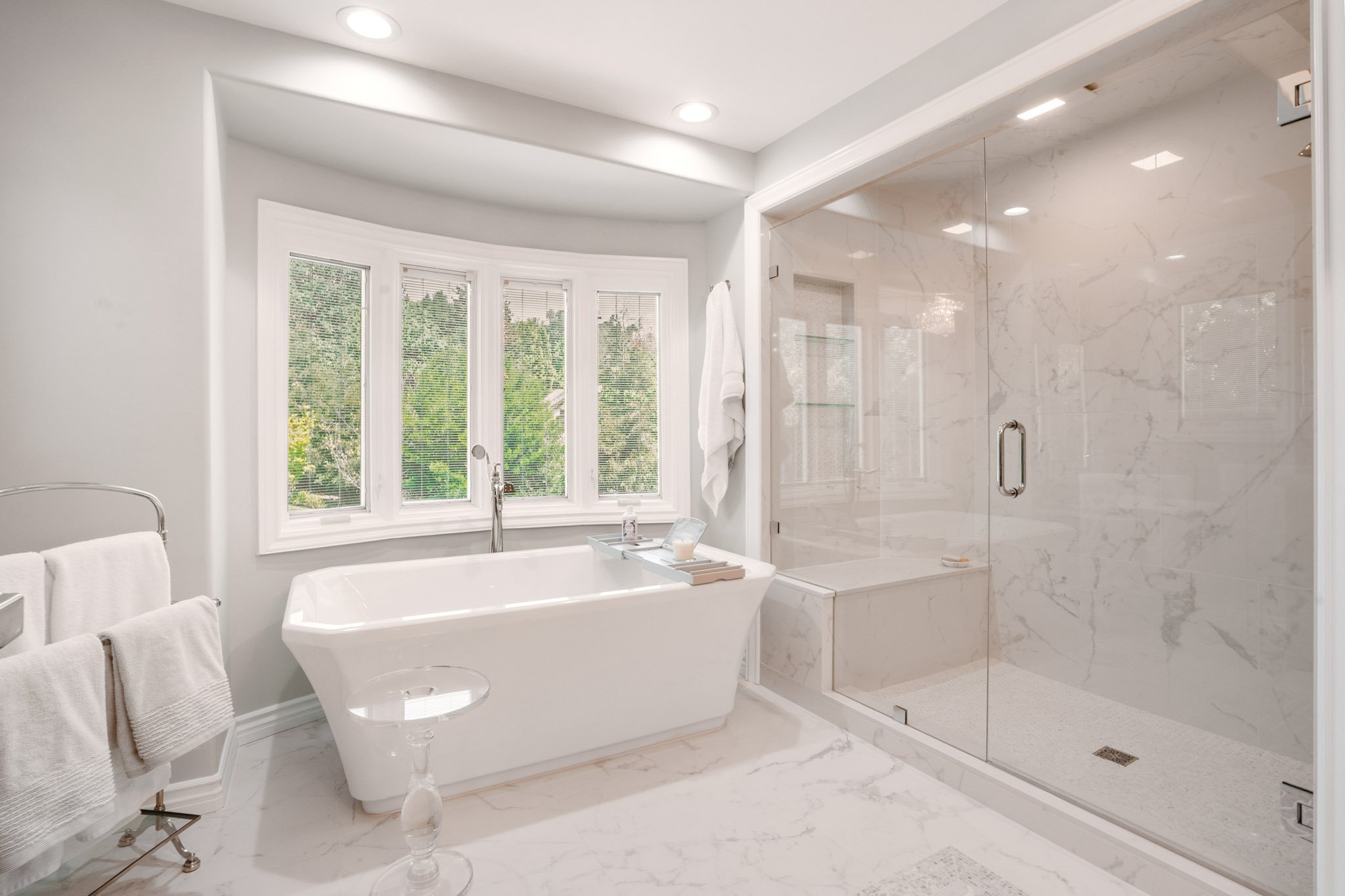 Bright and modern bathroom with a white freestanding bathtub beneath three large windows with blinds, a marble shower stall with glass door, and white towels on a towel rack.