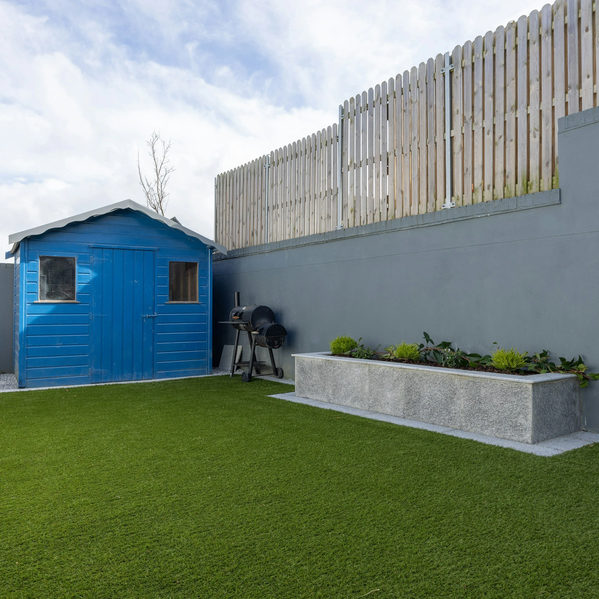A backyard scene with a well-maintained green lawn, a blue shed with windows, and a tall wood fence on top of a gray concrete wall. There are some small plants and shrubs in a planter in the foreground, and a grill is visible near the shed. The sky is partly cloudy with patches of blue.