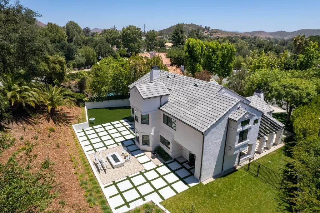 A large modern white house with a gray tiled roof, surrounded by lush green trees and a landscaped backyard with a checkered concrete and grass patio, outdoor seating, and a firepit.