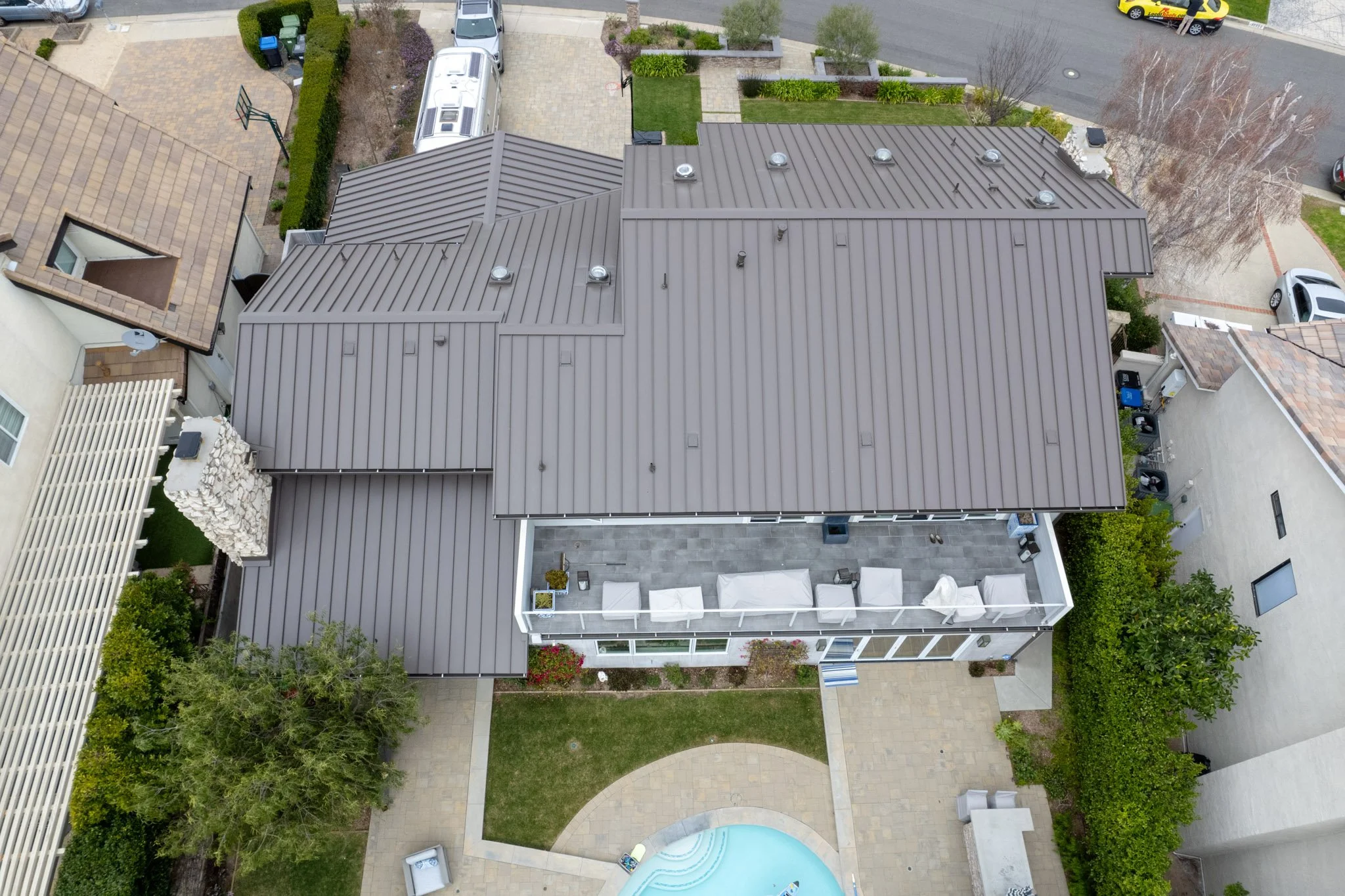 Aerial view of a residential backyard with a gray metal roof house, a swimming pool, patio furniture, a green lawn, trees, and surrounding streets with parked cars.