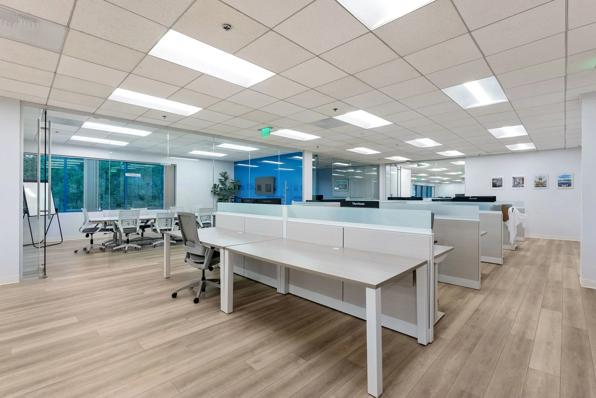 Open office space with multiple white desks and chairs, glass walls, and ceiling lights.
