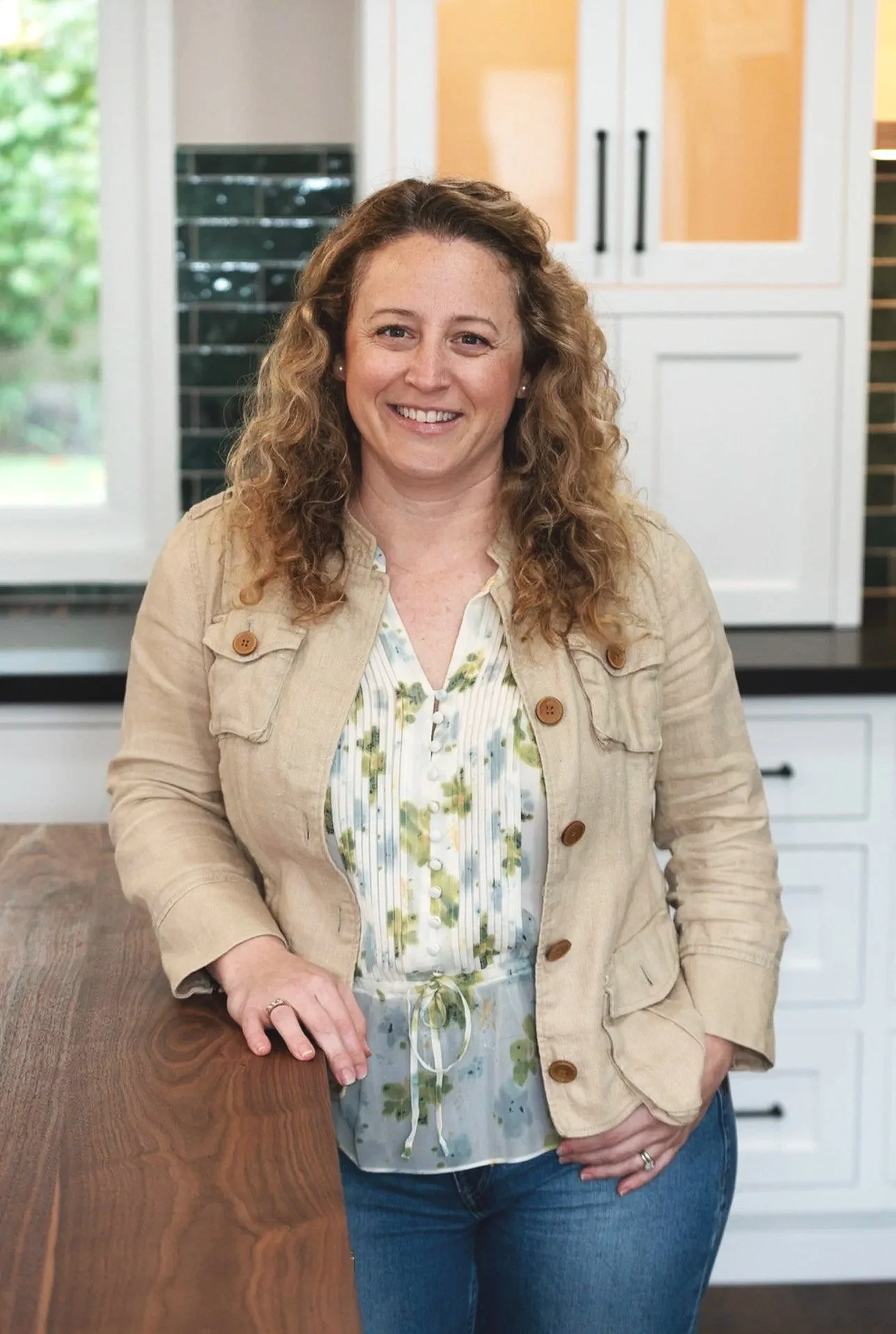 A woman with curly hair smiling, standing in a kitchen with white cabinets and a window showing greenery outside.