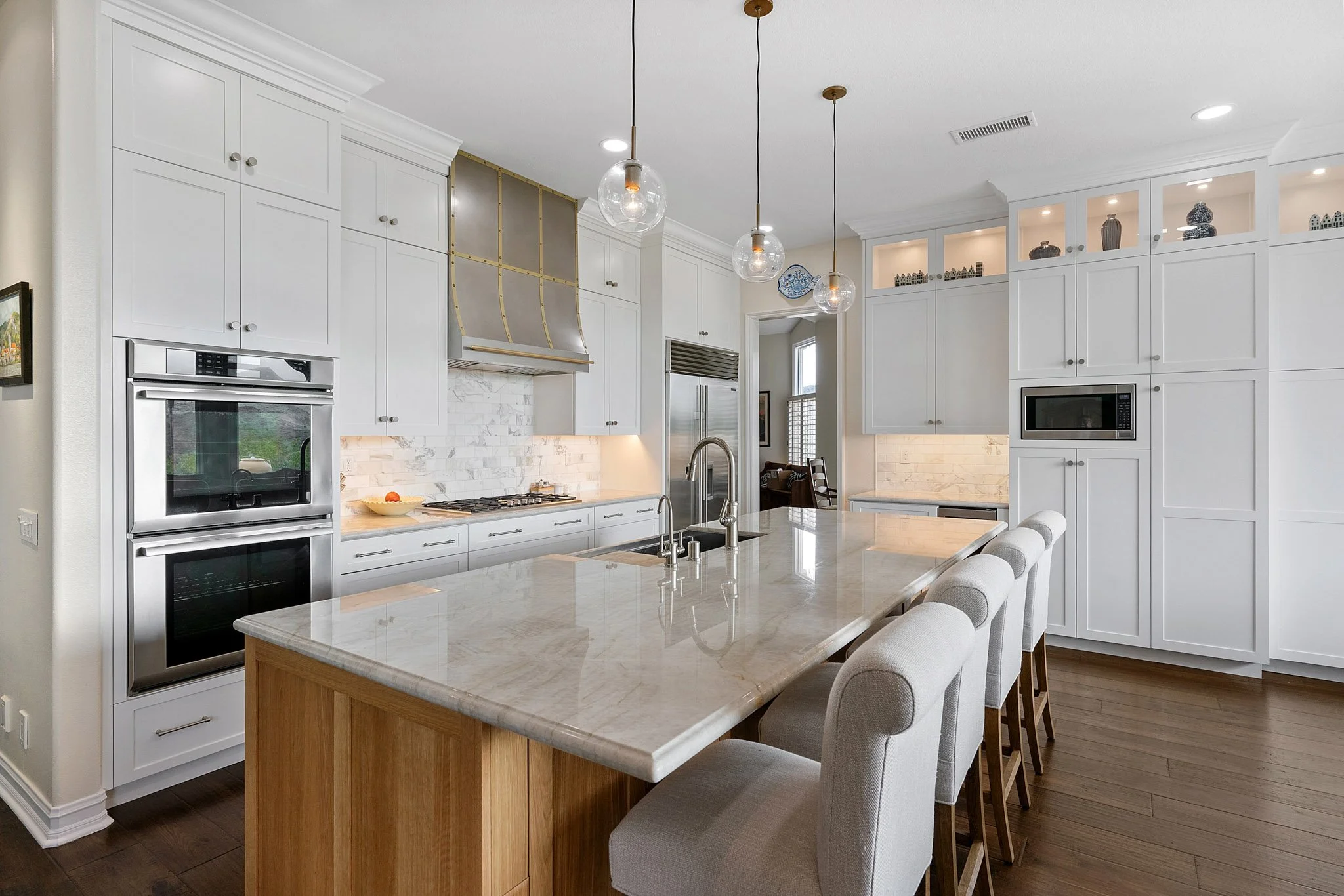 Modern white kitchen with an island and bar stools, built-in double oven, microwave, and glass pendant lights.