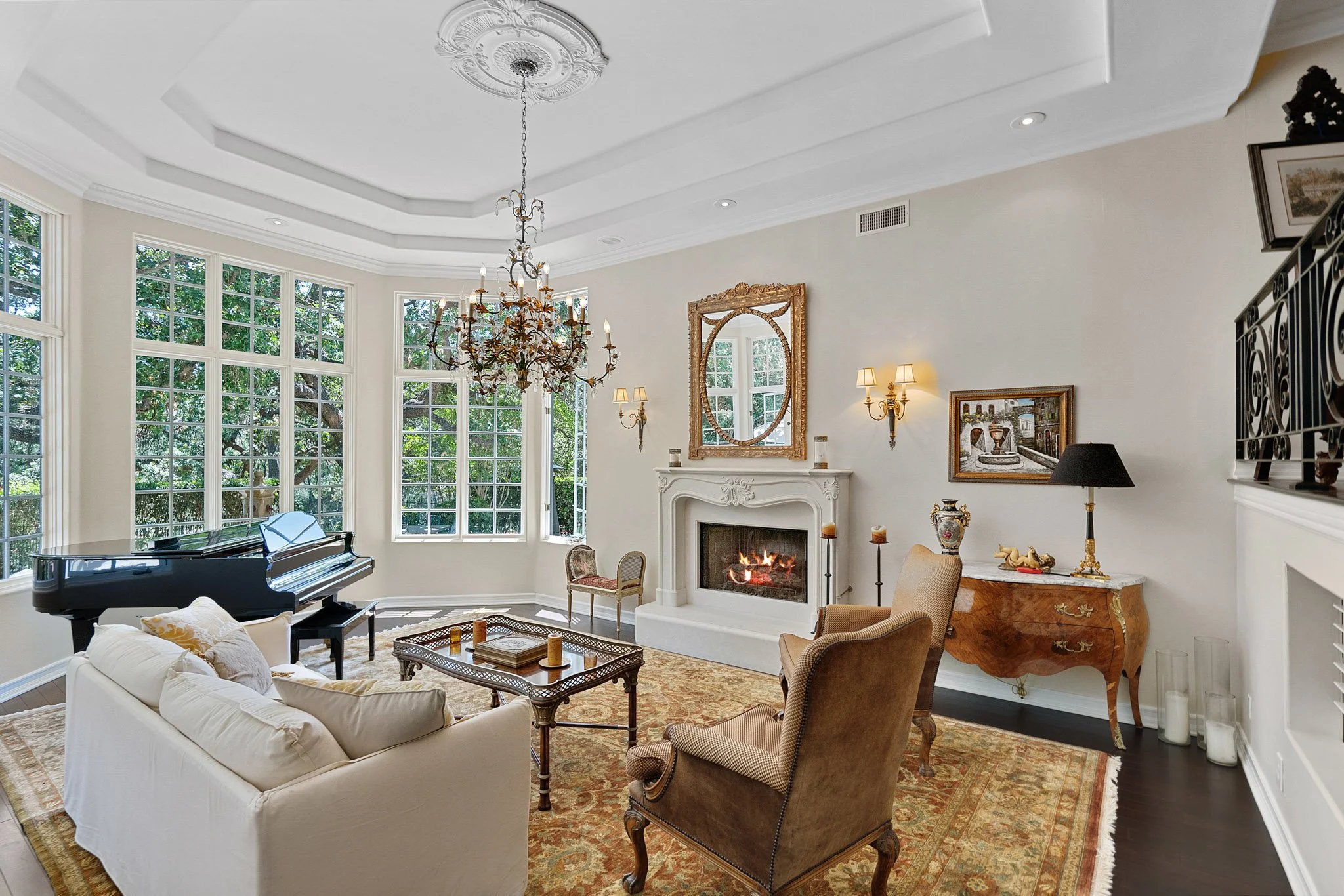 Elegant living room with large windows, a chandelier, a fireplace, a sofa, armchairs, a grand piano, artwork, and decorative items.