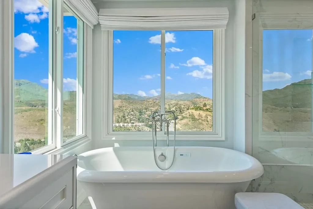 A white bathtub with a chrome faucet is positioned in front of large windows showing a scenic landscape with hills and a partly cloudy sky.