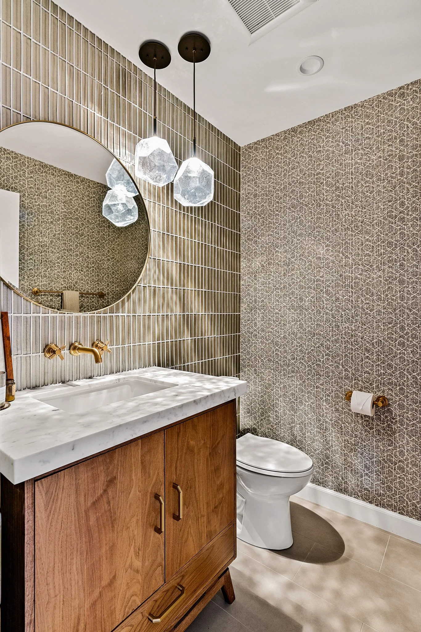 A bathroom featuring a wooden vanity with a white marble countertop, a round mirror, gold fixtures, a toilet, a textured tile wall, and hanging pendant lights with geometric glass shades.