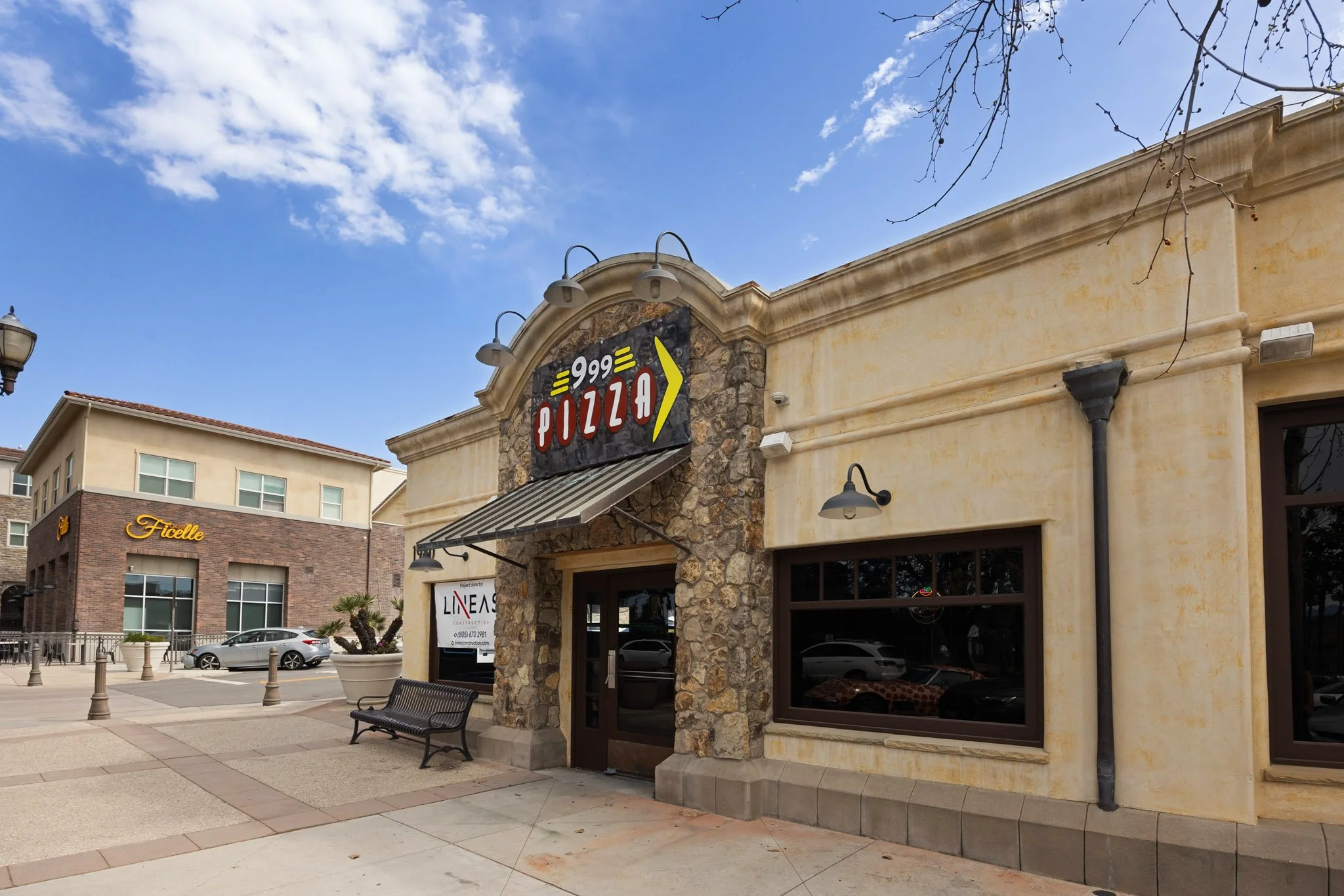 The image shows a shopping plaza with a pizza restaurant sign on a building with beige walls and a stone facade. There are parked cars and a few benches in the front, and a clear sky with some clouds above.