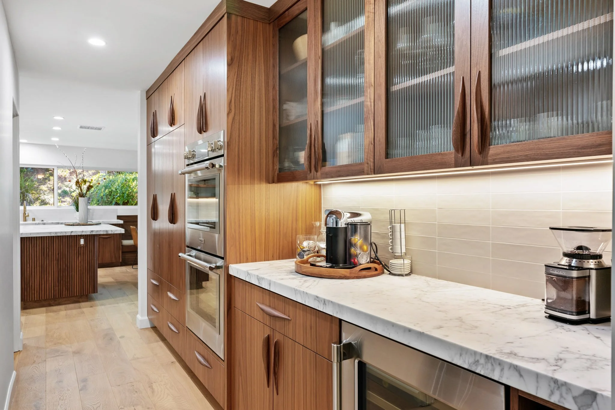 A modern kitchen with wooden cabinets, a white marble countertop, and various small appliances on the counter including a coffee maker and toaster. The kitchen has light wood flooring and a tile backsplash. In the background, there is a dining area with a window and a view of greenery outside.