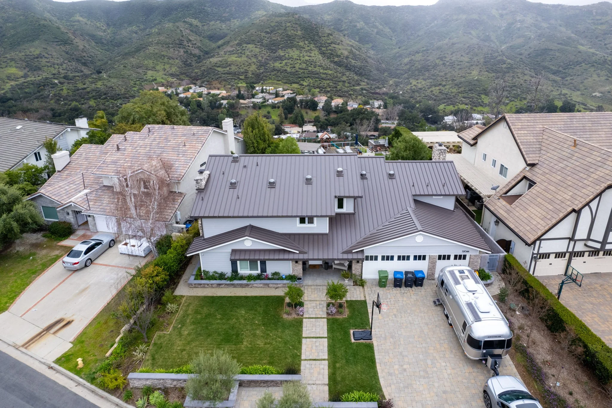 Aerial view of suburban neighborhood with houses, garages, parked cars, and green lawns, set against rolling green hills and mountains in the background.