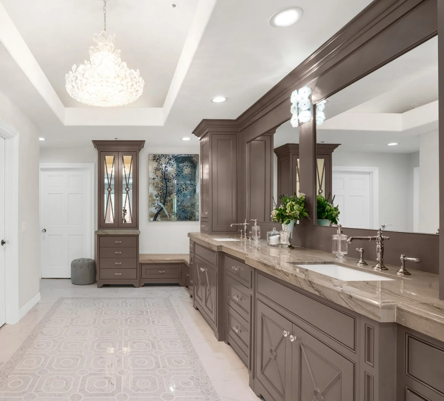 Elegant bathroom with taupe cabinetry, marble countertops, large mirror, floral arrangement, built-in cabinet, chandelier, and artwork on white walls.