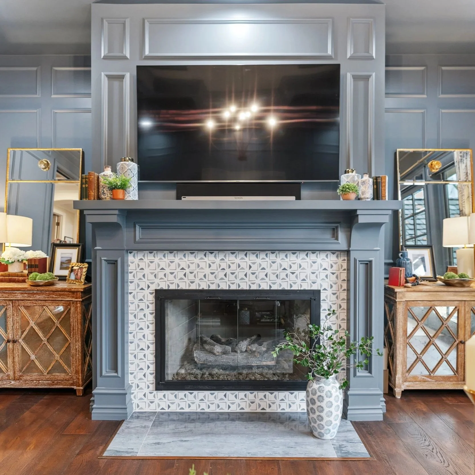 A living room fireplace with a flat-screen TV mounted above it, decorated with small plants, books, and decorative bottles on the mantel. To the sides are matching wooden cabinets with lattice wood fronts. There are framed photos and small decor items on and around the cabinets. The floor has wood planks and the fireplace is framed with blue molding and tile.