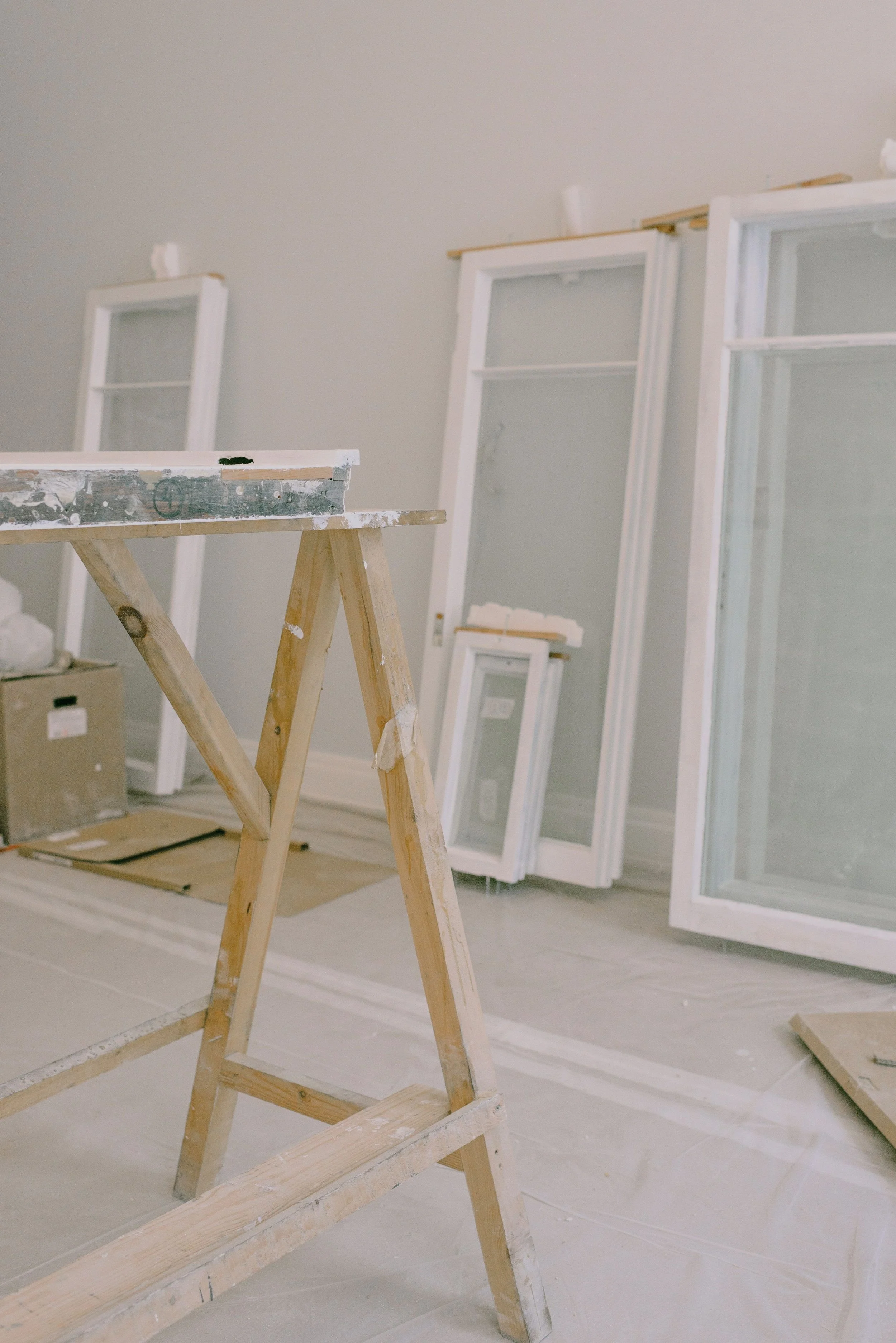 Interior of a room under construction with a wooden ladder and white window frames leaning against the wall.