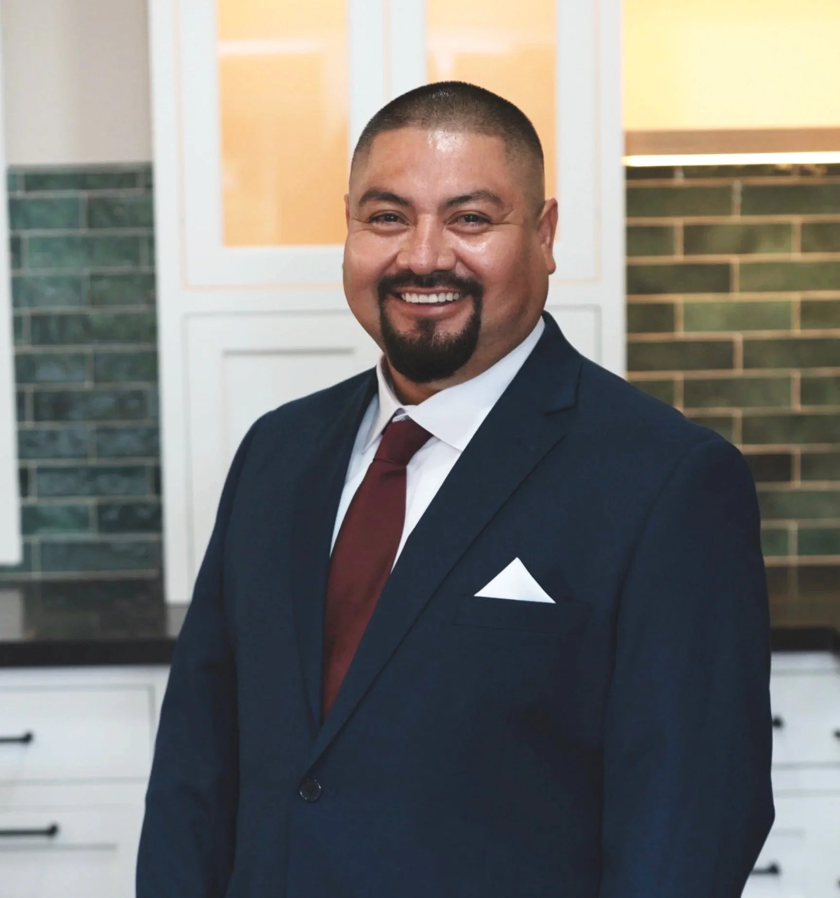 Close-up of a smiling man in a dark suit, white shirt, maroon tie, and a white pocket square, standing in front of a background with white cabinets and green tile walls.