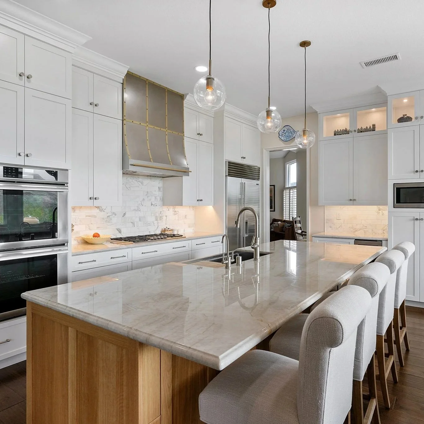 Modern kitchen with white cabinets, marble backsplash, and stainless steel appliances. A large marble island with seating, pendant lights above, and a visible adjacent living room.
