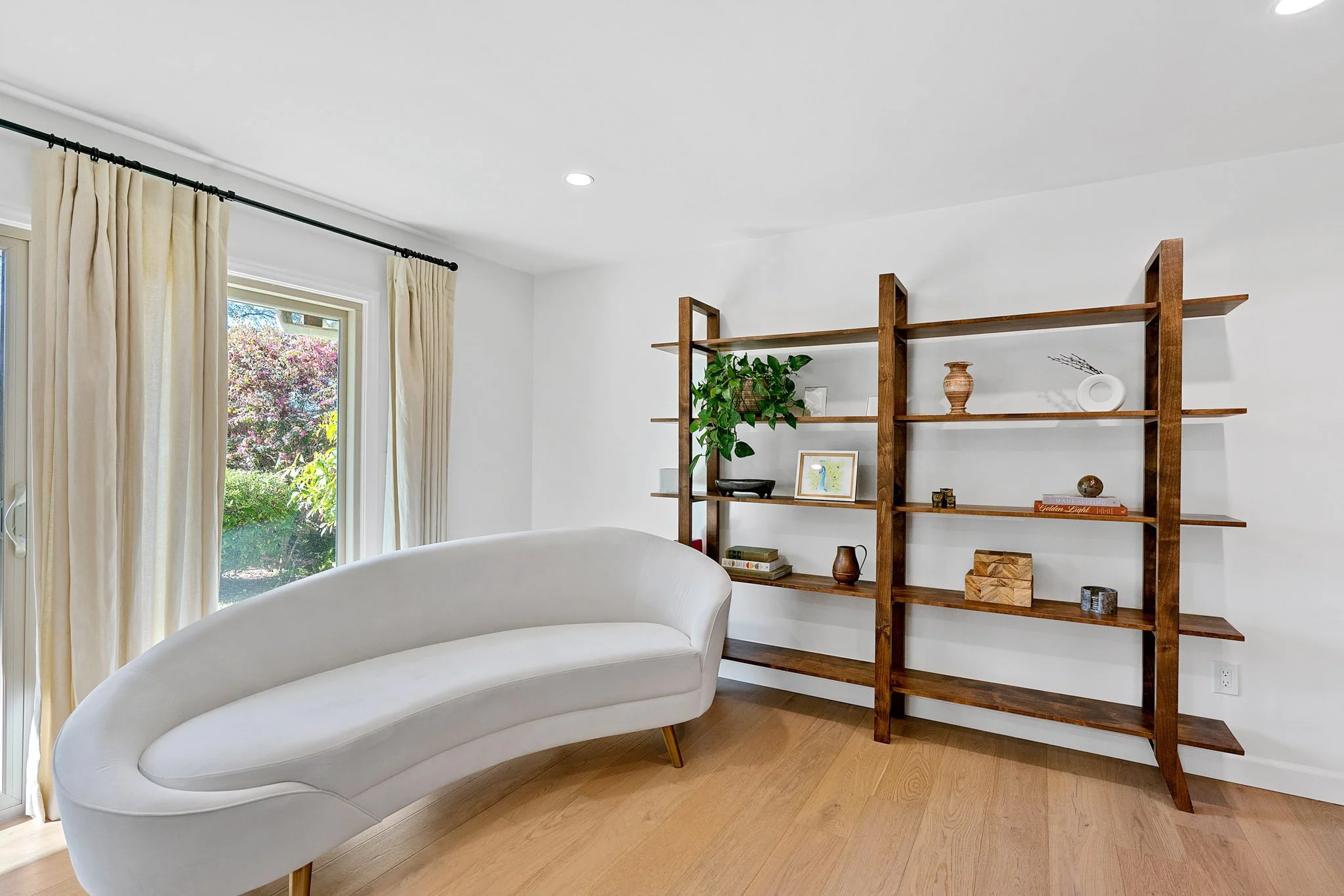 A living room with cream curtains over windows showing outdoor trees and flowers, a modern white curved sofa, and a wooden bookcase with decorative items.