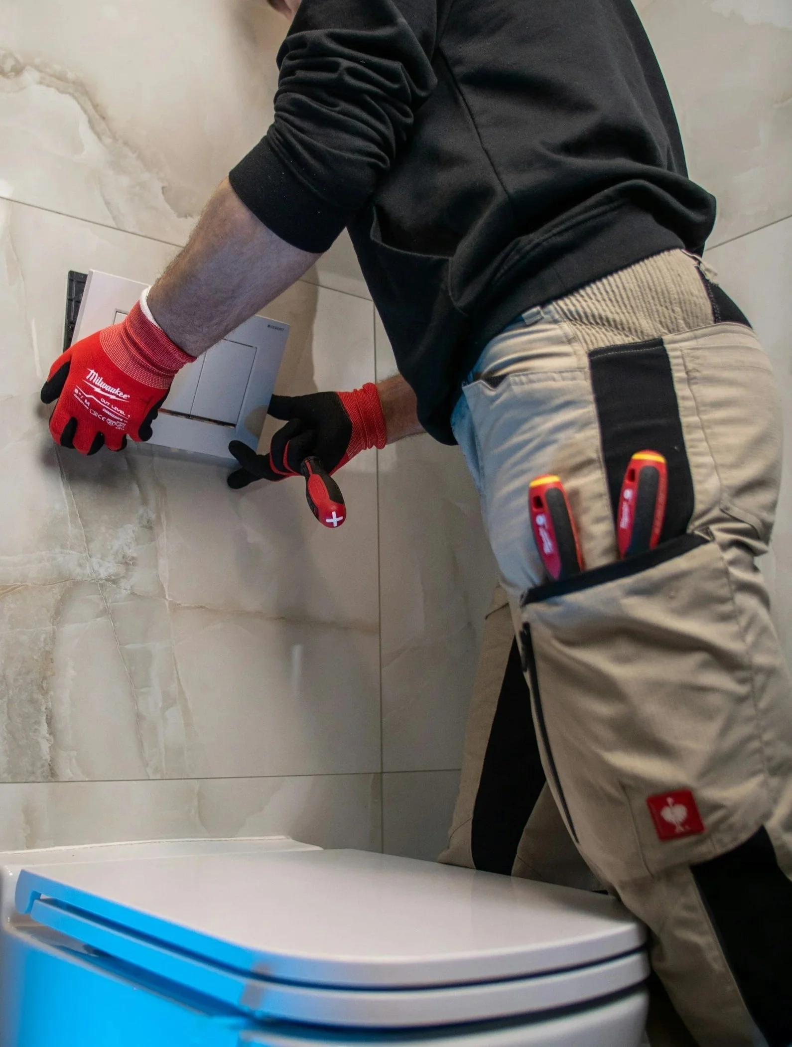 Person installing or fixing a white bathroom toilet with beige tiles on the wall, wearing a black long sleeve shirt, beige pants, red work gloves, and using a screwdriver.