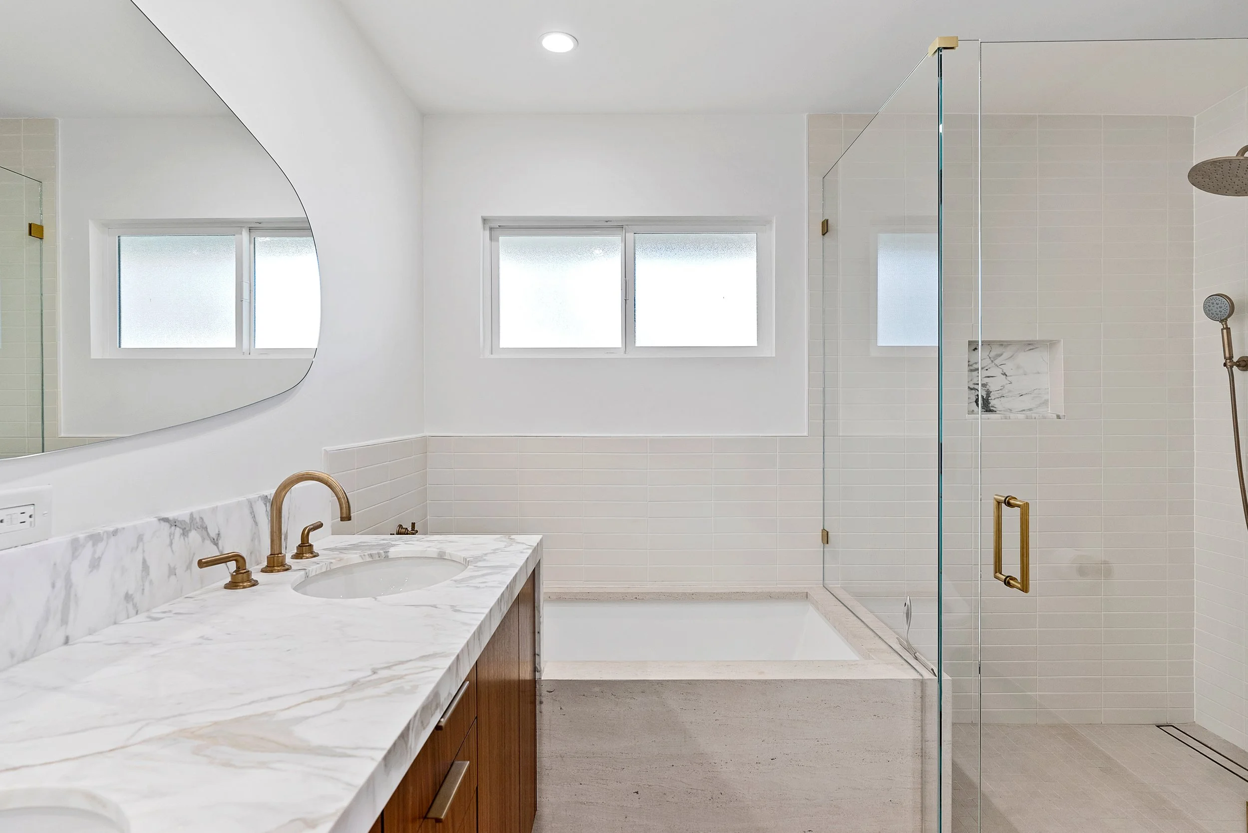 Modern bathroom with white tiled walls, a marble countertop with a sink and bronze fixtures, a large mirror, and a glass-enclosed shower with a rain showerhead.