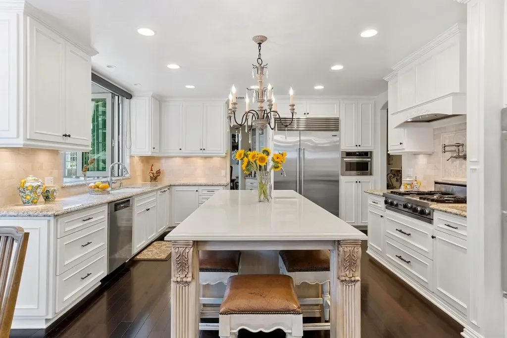 Bright white kitchen with large central island, sunflower centerpiece, stainless steel refrigerator, built-in oven, gas cooktop, and chandelier with candles, dark hardwood floors, and natural light from window.