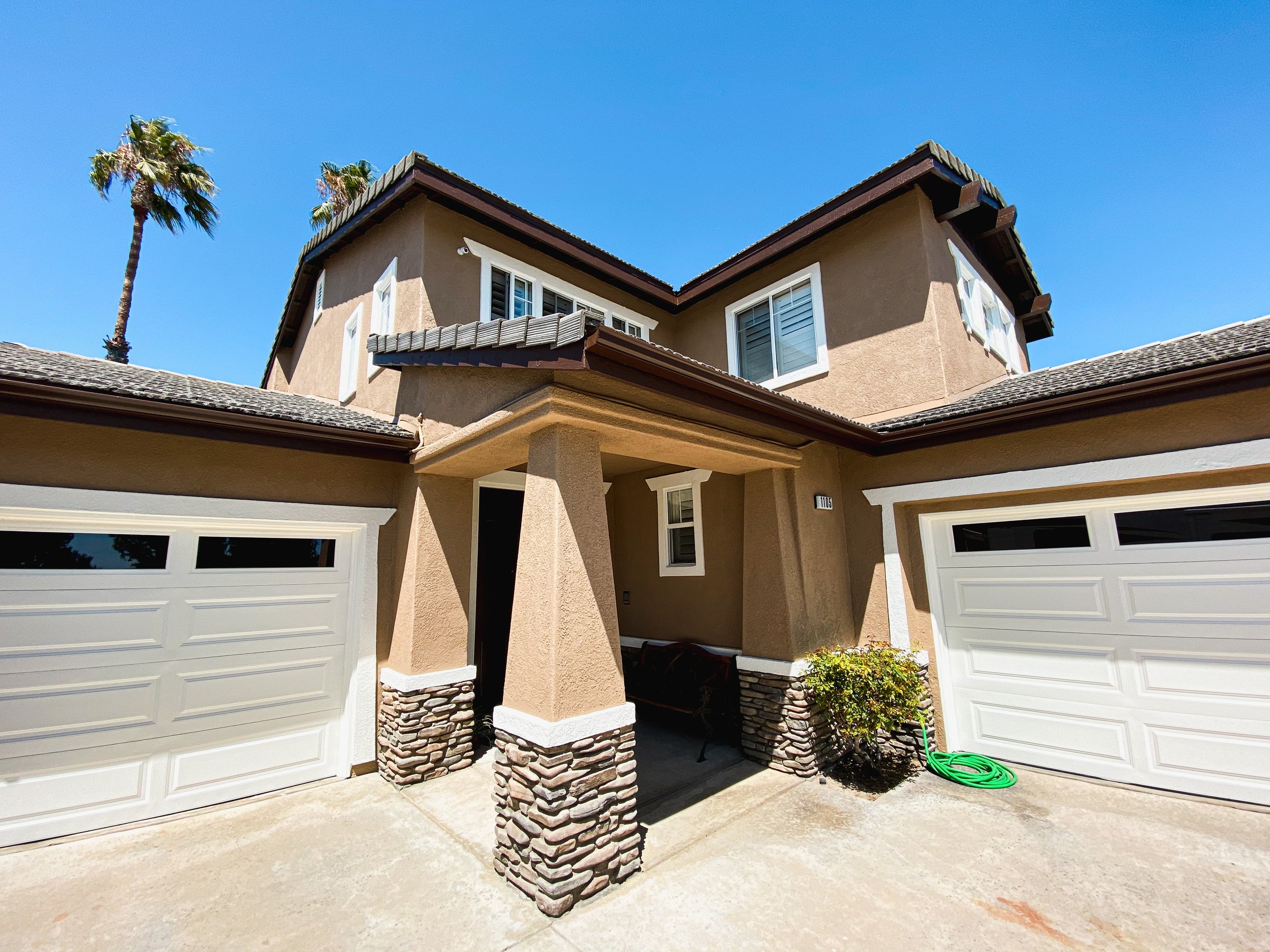 Front view of a two-story house with beige stucco exterior, brown roof, white trim, double garage doors, and palm trees against a clear blue sky.