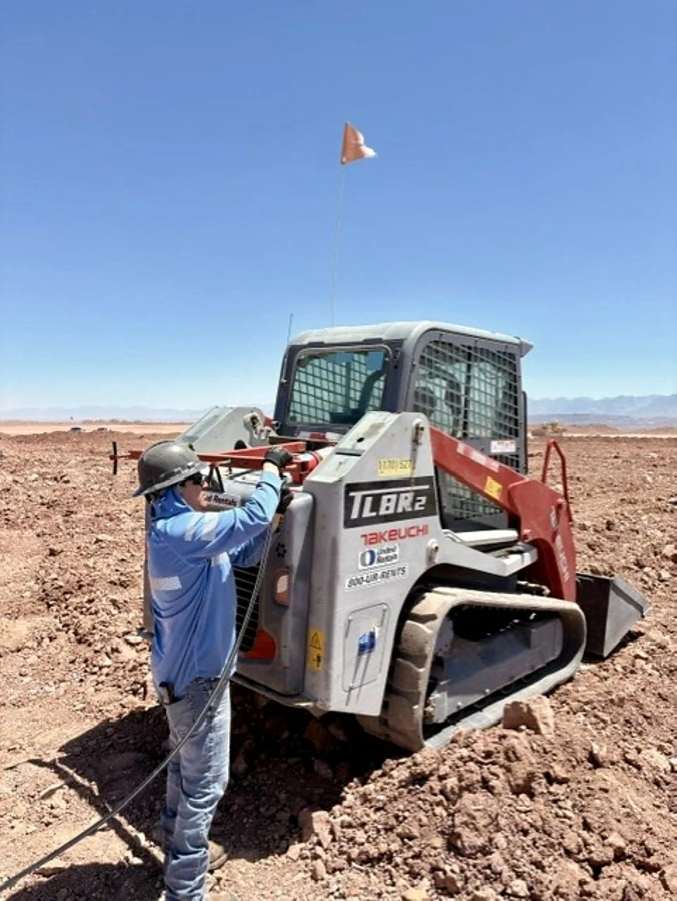 A construction worker in a blue shirt and helmet operates a small bulldozer in a desert landscape, with a blue sky and distant mountains.