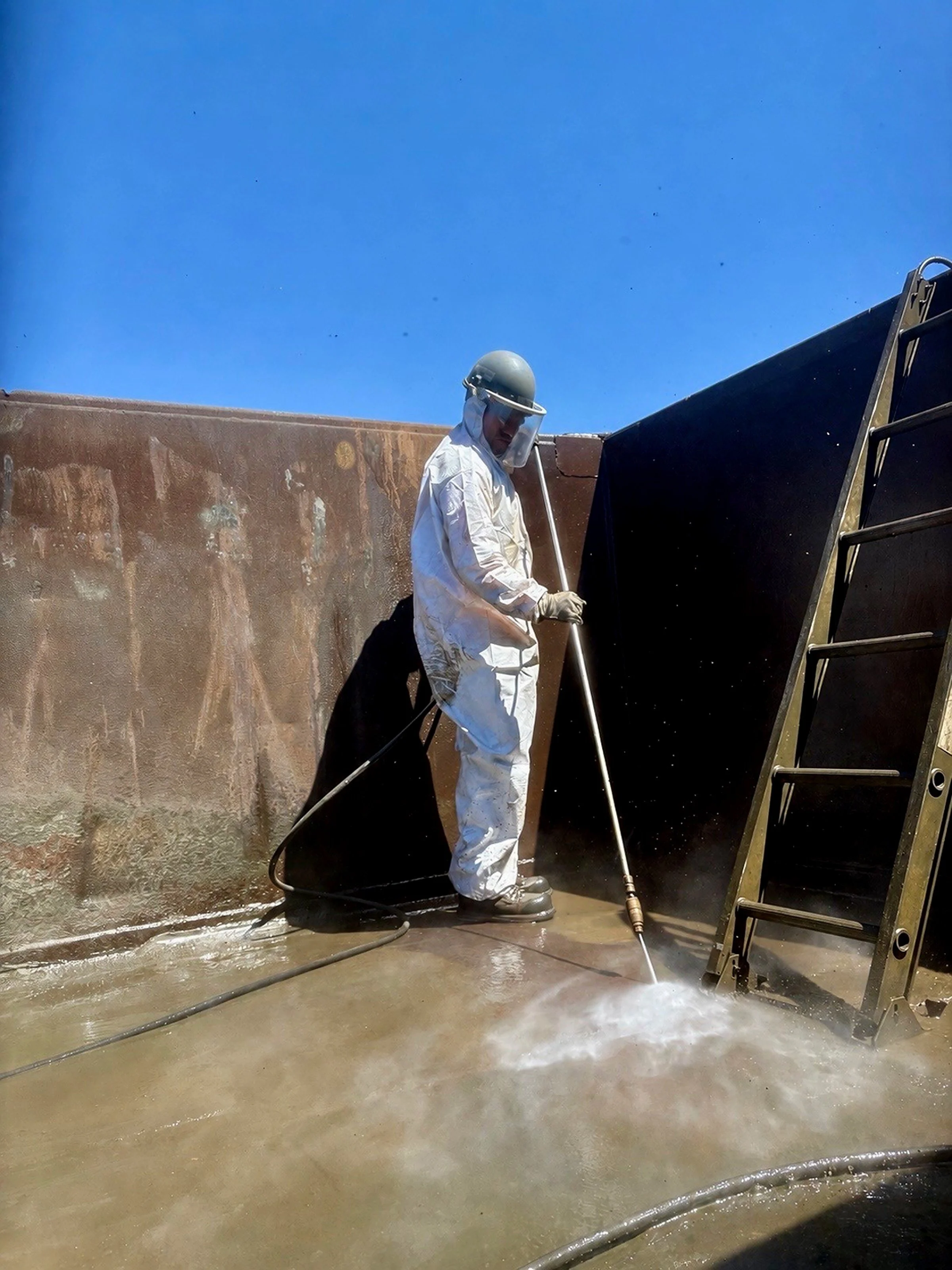 A Prime Industrial employee in Northern Nevada wearing a white protective suit, helmet, and face shield power washing a mine equipment with water spray, next to a ladder and a large dark object, against a blue sky.