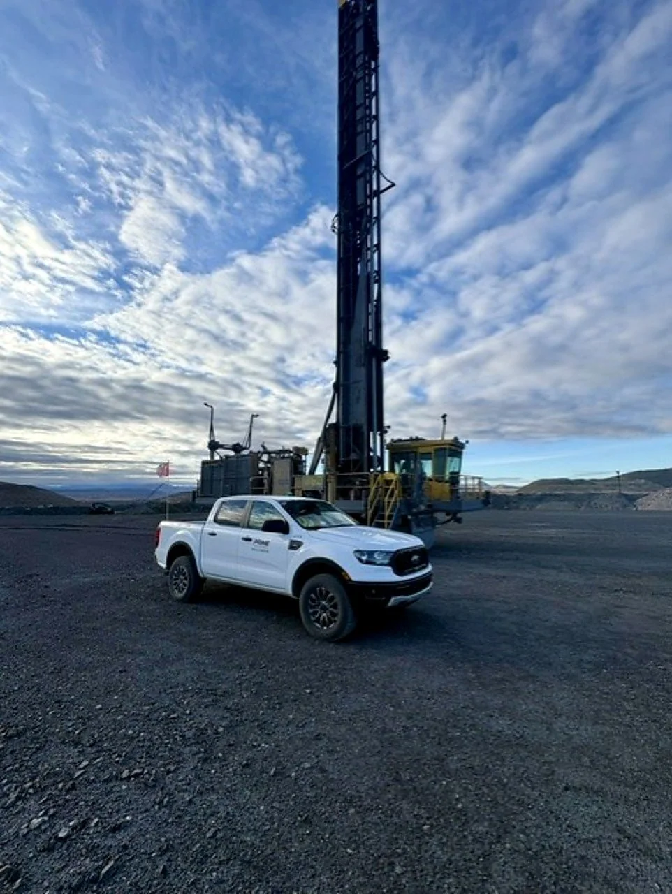 A white Ford Prime Industrial pickup truck parked on gravel in front of a large drilling rig in a rugged outdoor landscape with a partly cloudy sky.