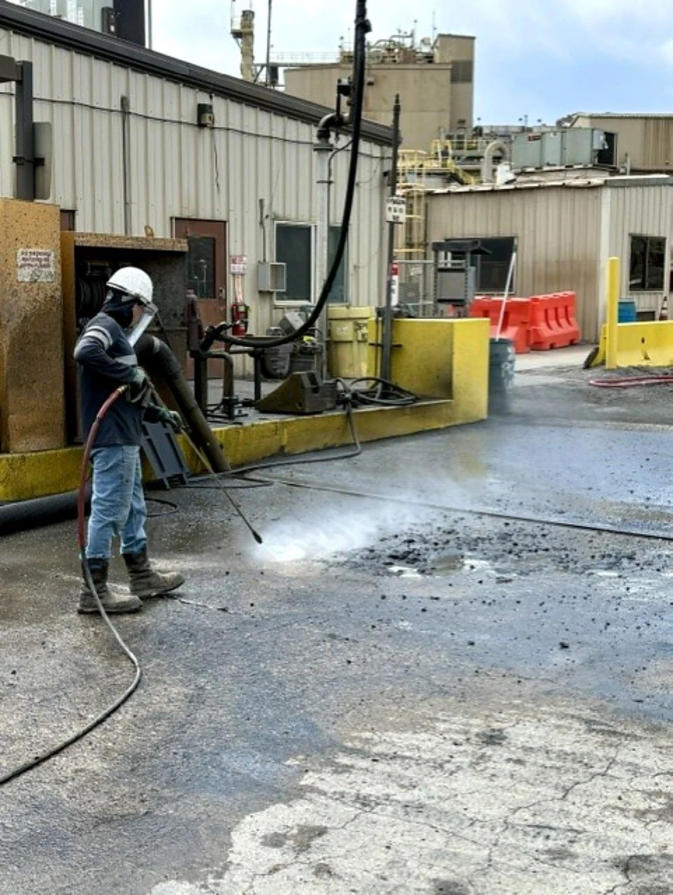 Worker using a jackhammer to break asphalt outdoors near industrial buildings, with safety gear including a helmet and face shield.
