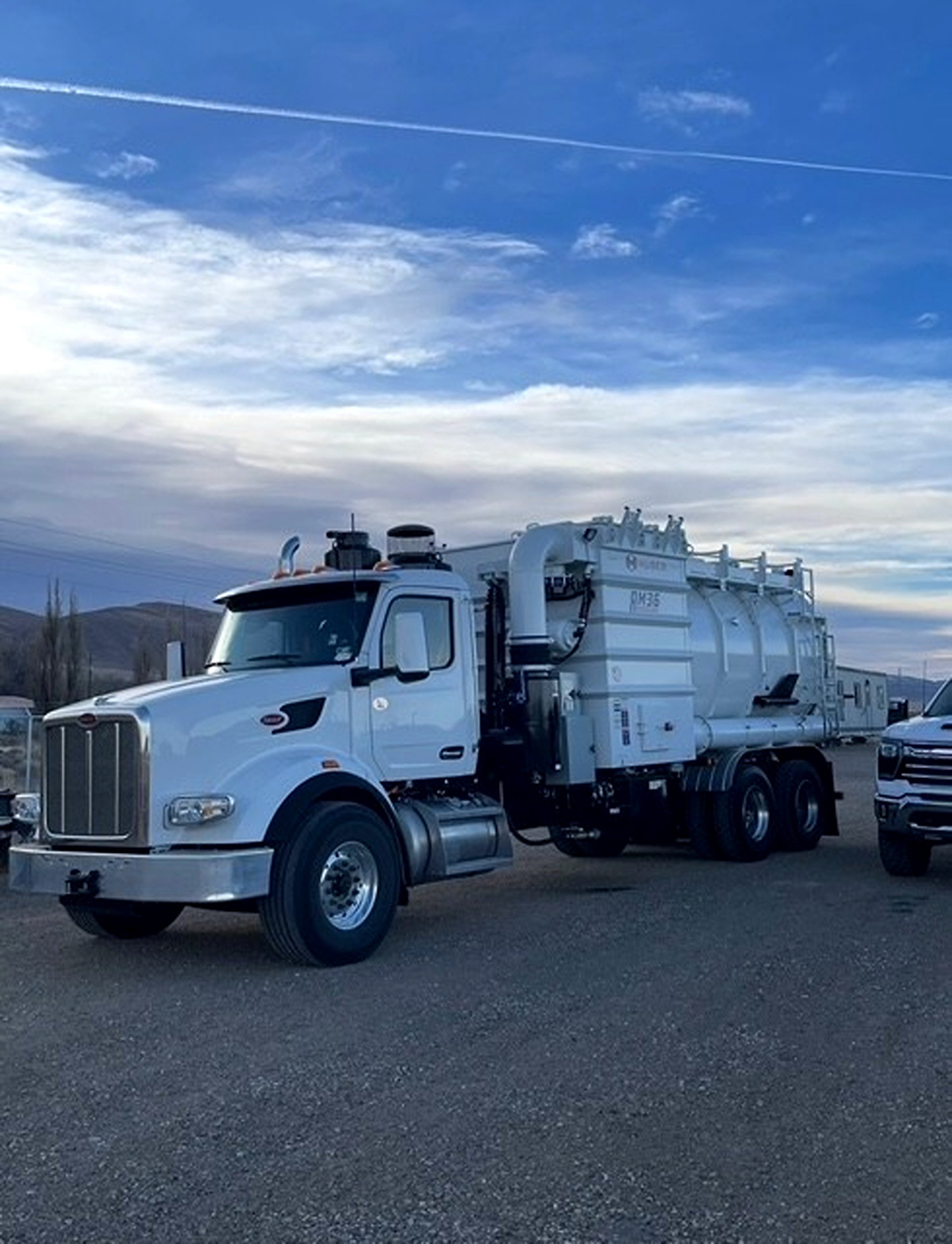 White utility truck equipped with specialized equipment, parked outdoors on a gravel lot with a cloudy sky overhead.